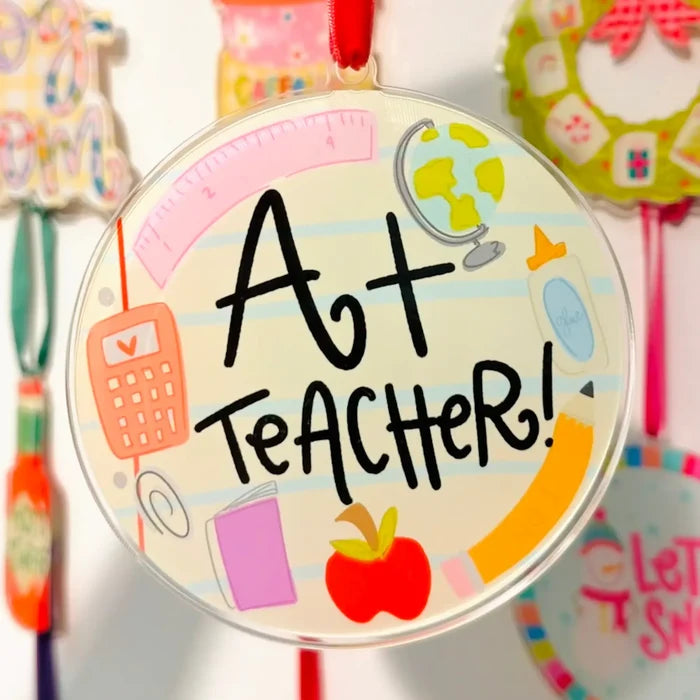 A round hanging ornament with a red ribbon loop at the top, featuring the words “Art Teacher!” in black script over a school-themed illustration with an apple, ruler, calculator, pencil, globe, and other colorful school supplies.