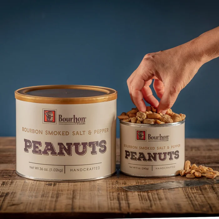 Two cans of Bourbon Smoked Salt & Pepper Peanuts with a hand reaching into one can, on a wooden surface with a blue background.