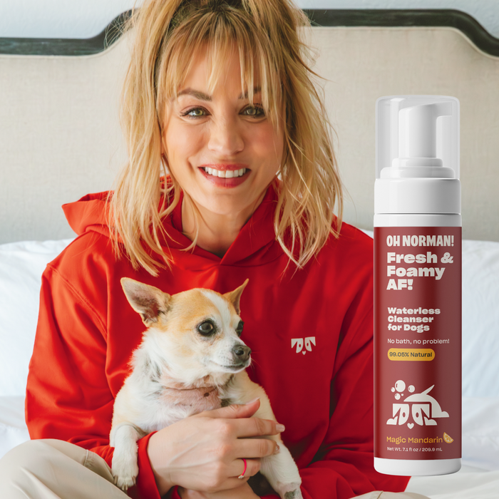 Kaley Cuoco in a red hoodie holding a small dog on a bed with a bottle labeled Oh Norman Fresh and Foamy AF waterless cleanser visible at right