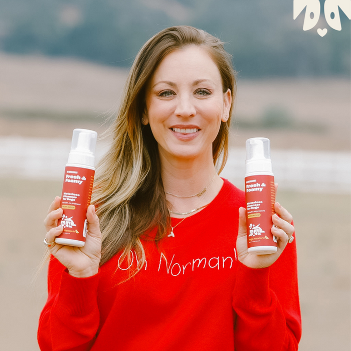 Kaley Cuoco  in red sweater holding two bottles labeled 'Fresh & Foamy Waterless Cleanser for Dogs no bath, no problem rojo mandarin'